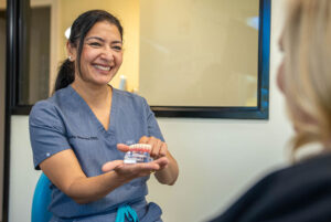 A smiling dental professional in scrubs holds a dental model, explaining tooth care to a seated patient in an office setting.