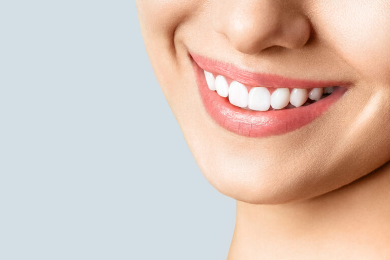A close-up of a smiling persons mouth, showing white teeth and smooth skin against a light blue background.