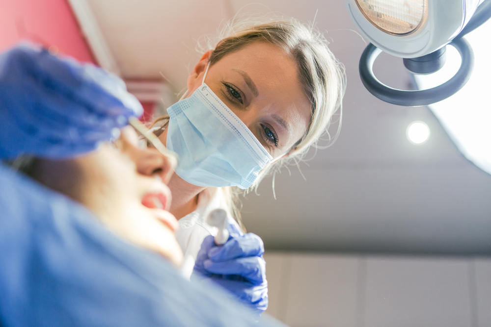 A dentist wearing a face mask and blue gloves examines a patients mouth under a bright examination light in a dental clinic.