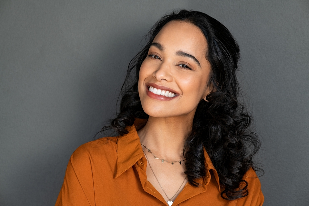 Woman with long, curly dark hair smiling at the camera, wearing an orange blouse and layered necklaces, standing against a neutral gray background.