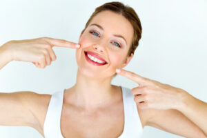A smiling woman with blue eyes points to her mouth with both index fingers. She is wearing a white top and has her brown hair tied back, standing against a plain white background.