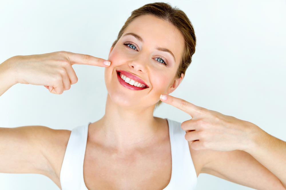A smiling woman with blue eyes points to her mouth with both index fingers. She is wearing a white top and has her brown hair tied back, standing against a plain white background.
